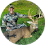 A person in camouflage clothing kneels on grass, smiling and holding the antlers of a large deer lying on the ground. The scene is outdoors in a grassy area, and the person appears to be posing after a successful hunt.