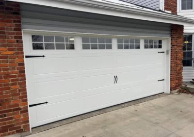 A white double garage door with decorative black handles and hinges, featuring four rectangular windows at the top. The door is set into a red brick house with concrete driveway in front.