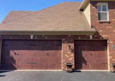 A brick house with two large, brown, wooden garage doors, a sloped shingle roof, a tan exterior wall, a wall-mounted lantern, and a planter in front of the garages. The sky is clear and blue.
