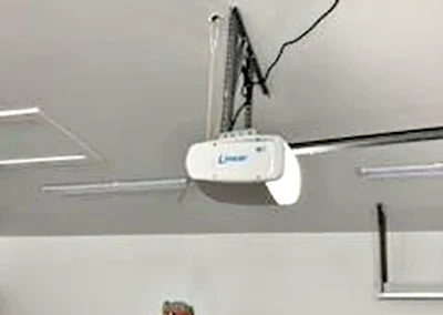 A garage ceiling with a mounted automatic garage door opener, visible wiring, and a red ladder positioned underneath. The garage door tracks and part of an open garage door are also visible.