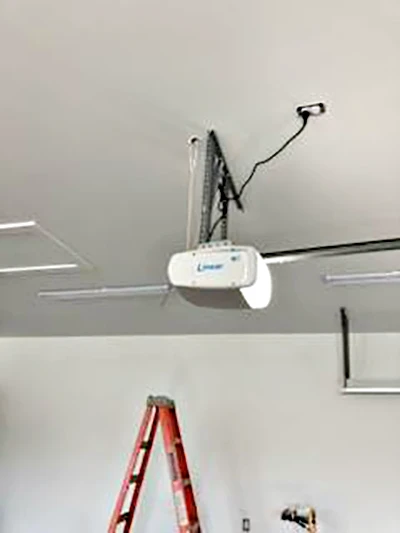 A garage ceiling with a mounted automatic garage door opener, visible wiring, and a red ladder positioned underneath. The garage door tracks and part of an open garage door are also visible.