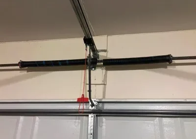 A close-up view of a garage door torsion spring system mounted above a closed white garage door, with a red emergency release cord hanging down.