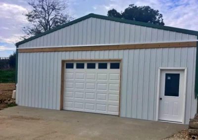 A white metal building with a single garage door and a separate white entrance door, set on a concrete driveway, with trees and a partly cloudy sky in the background.