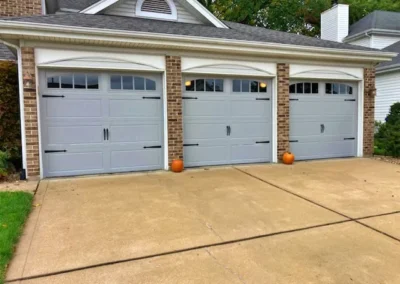 A three-car garage with light gray doors; each door has black decorative hardware. Three small pumpkins are placed in front of the doors, one by each. The driveway is clean, and the house has brick accents.