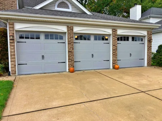 A three-car garage with light gray doors; each door has black decorative hardware. Three small pumpkins are placed in front of the doors, one by each. The driveway is clean, and the house has brick accents.