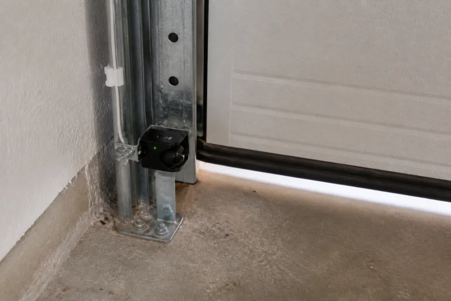 A close-up of a garage door sensor mounted near the bottom metal track and concrete floor, with the garage door partially open, showing a small gap underneath—a reminder to regularly check your system’s components when considering how long do garage door openers last.