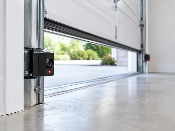 Close-up of a garage door safety sensor with a red light on, mounted near the bottom of the door track. The garage door is partially open, and daylight is visible outside.