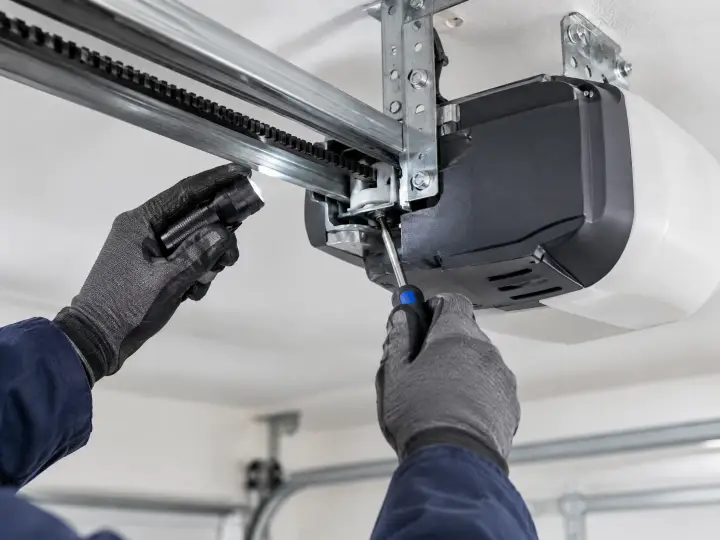 A person wearing gray gloves uses a screwdriver to repair or install a garage door opener mechanism attached to the ceiling.