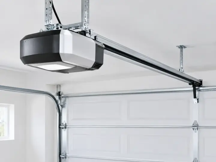 A close-up of a modern garage door opener mounted on the ceiling, with a metal track leading to a closed white garage door in a well-lit garage.