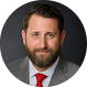 A man with brown hair and a beard, wearing a gray suit, white shirt, and red tie, poses for a professional headshot against a dark background.